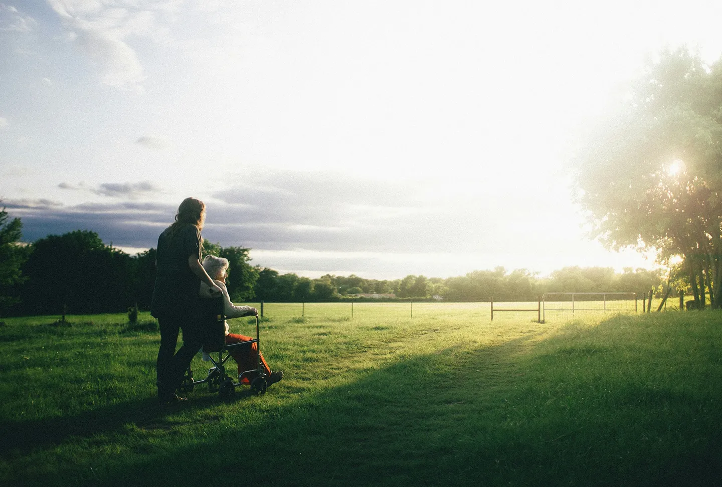 A caregiver walking beside an older adult resident through a sunlit field at golden hour